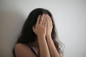 An adult woman with long brunette hair covers her face with her hands against a white wall, having issues with vitamin b1 thiamine deficiency